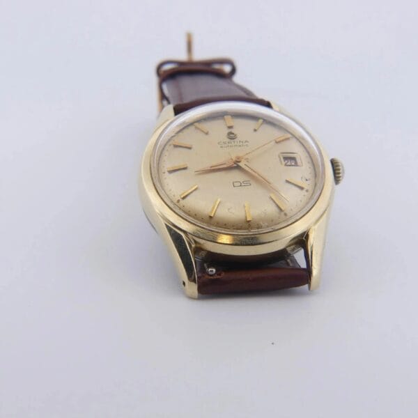 A 1959 Certina DS Automatic 1st Gen. wristwatch featuring a gold cap and brown leather strap, gold hour markers, a date window at the 3 oclock position, and the brand name displayed on a plain white background.