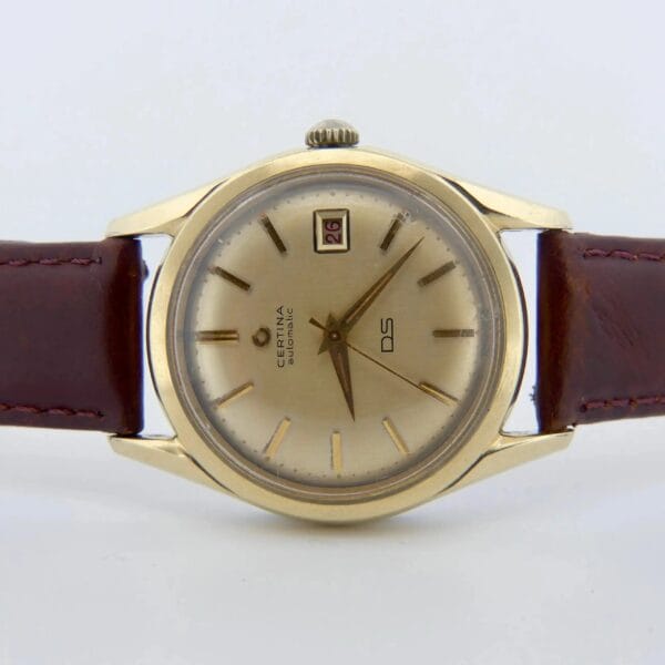 The 1959 Certina DS Automatic 1st Gen. 14k Gold Cap features a gold-toned round face, brown leather strap, simple hour markers, two hands, and a date window at 3 oclock. Certina DS is printed on the dial.