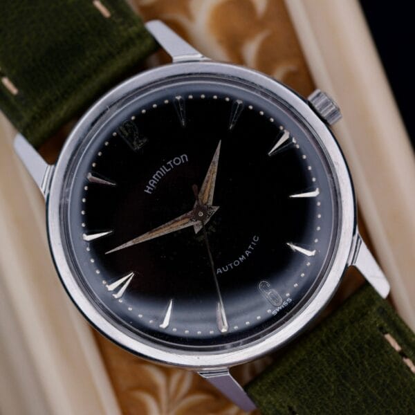 A close-up of a 1960 Hamilton Thin-o-Matic T-501 Micro-Rotor automatic wristwatch with a black dial, silver hour markers, and green fabric strap on a textured surface. The watch displays approximately 9:12.