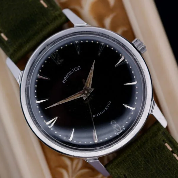A close-up of a 1960 Hamilton Thin-o-Matic T-501 Micro-Rotor automatic wristwatch with a black dial, silver hour markers, and green fabric strap on a textured surface. The watch displays approximately 9:12.