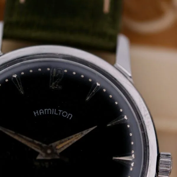 Close-up of a 1960 Hamilton Thin-o-Matic T-501 Micro-Rotor wristwatch with a black dial, silver hour markers, Hamilton on the face, showing 11:11. The watch features a green strap.