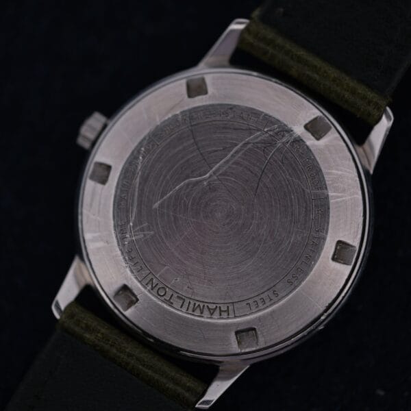Close-up of the back of a 1960 Hamilton Thin-o-Matic T-501 Micro-Rotor wristwatch with visible scratches and text around the edge, attached to a dark green leather strap.