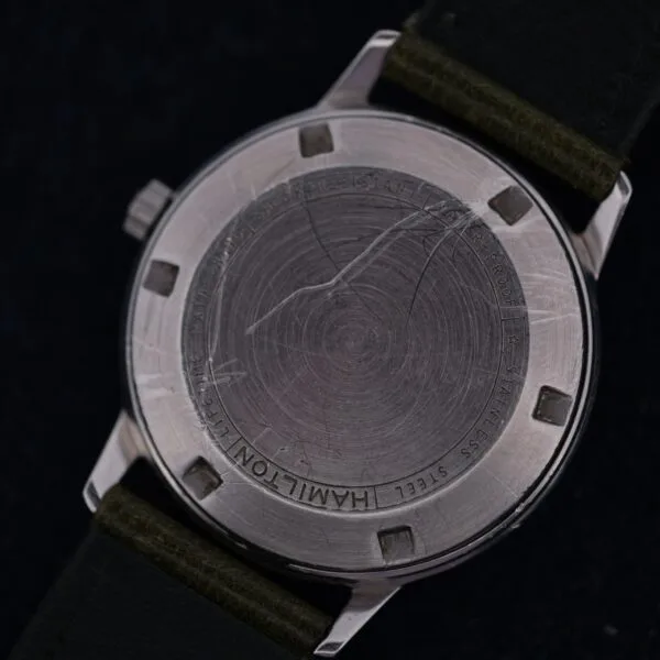 Close-up of the back of a 1960 Hamilton Thin-o-Matic T-501 Micro-Rotor wristwatch with visible scratches and text around the edge, attached to a dark green leather strap.