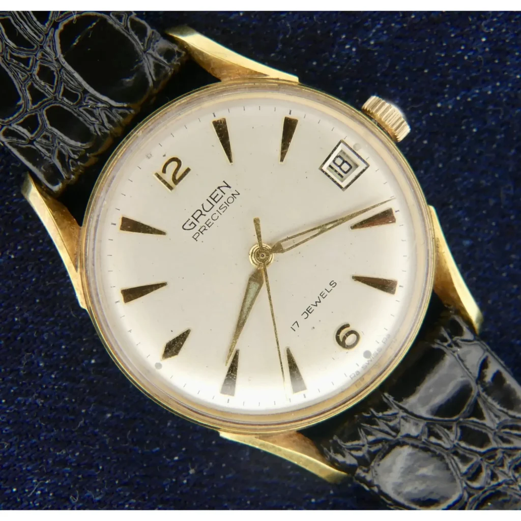 A 1960s Gruen Precision Bombay Lugs Cal. 510CA wristwatch with gold case, white dial, black indices, gold hands, date window at 3 o’clock, and “17 Jewels” text is shown on a black leather strap against a dark blue background.