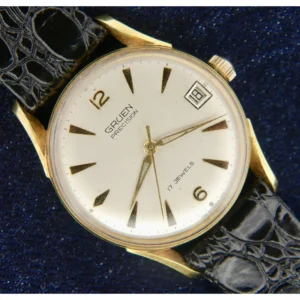 A 1960s Gruen Precision Bombay Lugs Cal. 510CA wristwatch with gold case, white dial, black indices, gold hands, date window at 3 o’clock, and “17 Jewels” text is shown on a black leather strap against a dark blue background.