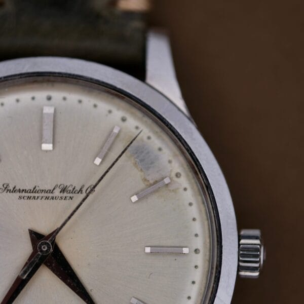 A close-up of a 1960s IWC Automatic Dress Watch Cal. 853 showing part of the white dial with silver hour markers, hands, crown, a visible scratch, and the text “International Watch Co SCHAFFHAUSEN.”.