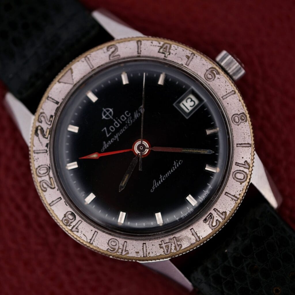 A vintage 1960s Zodiac Aerospace GMT Automatic Date wristwatch featuring a black face, date window, worn silver bezel, and black strap.