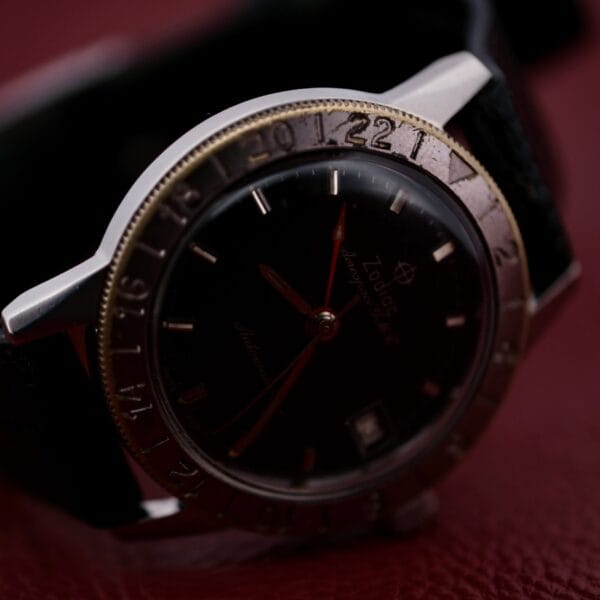 A close-up of the 1960s Zodiac Aerospace GMT Automatic Date, a black wristwatch with a metal bezel featuring 24-hour markers, red second hand, and leather strap, displayed on a textured dark red surface.