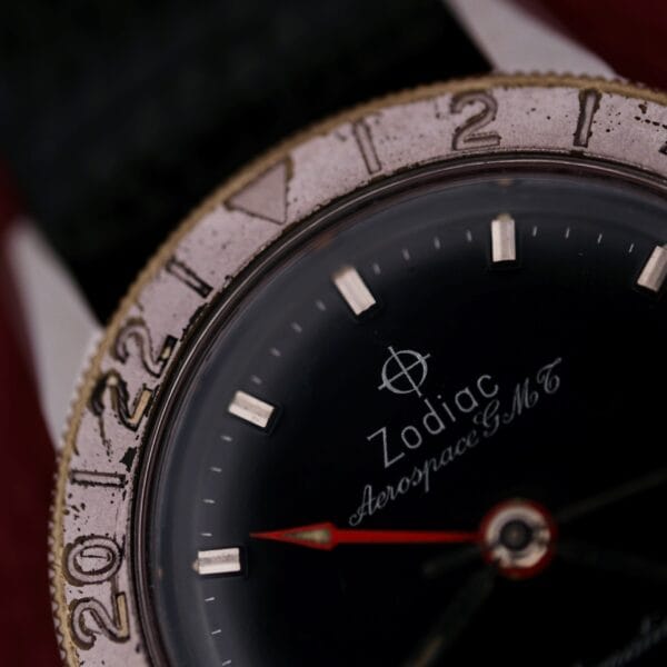 Close-up of a 1960s Zodiac Aerospace GMT Automatic Date watch, featuring a worn silver bezel with numbers, black dial, silver hour markers, red GMT hand, and white model text.