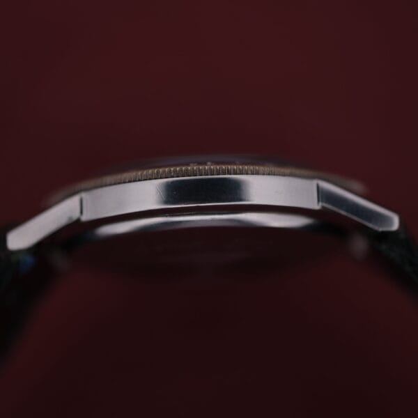 A close-up side view of the 1960s Zodiac Aerospace GMT Automatic Date shows its slim silver case, black strap ends on either side, set against a dark red background.