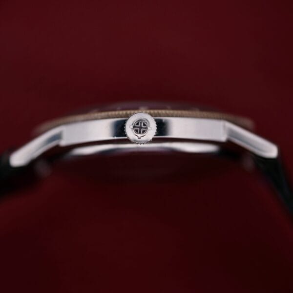 Close-up side view of the 1960s Zodiac Aerospace GMT Automatic Date in silver, set against a dark red background, highlighting the crown with its engraved pattern at the center.