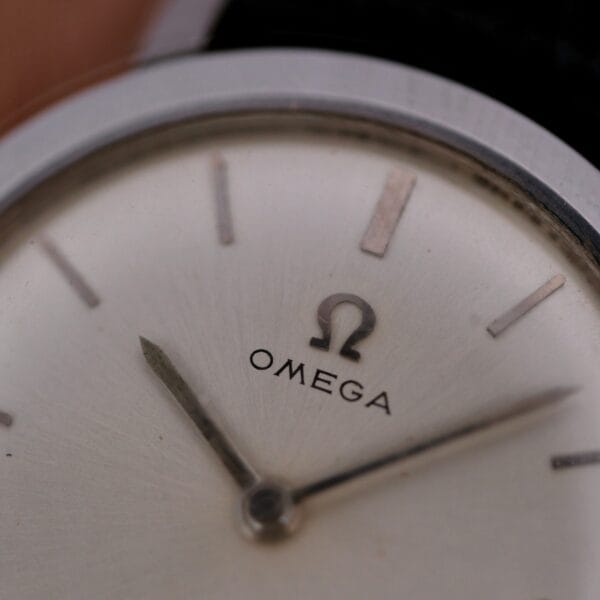 A close-up of the 1962 Omega Slim Dress Sunburst Dial shows its face with the Omega logo and brand name. The watch has minimalistic hour markers and hands on a light-colored background, with the edge of the case visible.