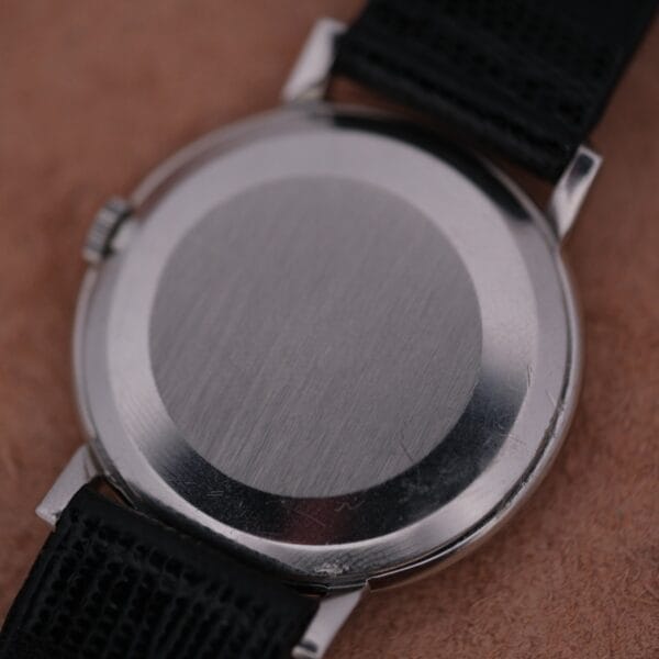 Close-up image of the 1962 Omega Slim Dress Sunburst Dial, featuring a polished metal case with a smooth back, black textured strap, and visible crown on a brown surface.