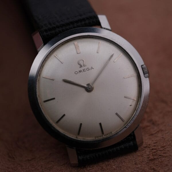 Close-up of a 1962 Omega Slim Dress Sunburst Dial watch, featuring a vintage minimalist design with a silver case, plain silver dial, thin black hour markers, two slim hands, and Swiss Made at the bottom. It includes a black leather strap.