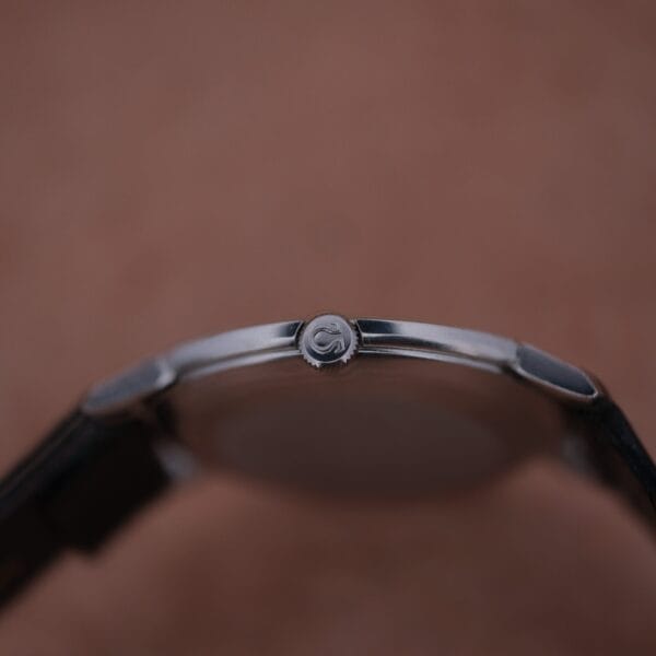 Close-up of the 1962 Omega Slim Dress Sunburst Dials side, highlighting the crown with a letter T logo. The watch features a metallic case and a black leather strap against a blurred beige background.