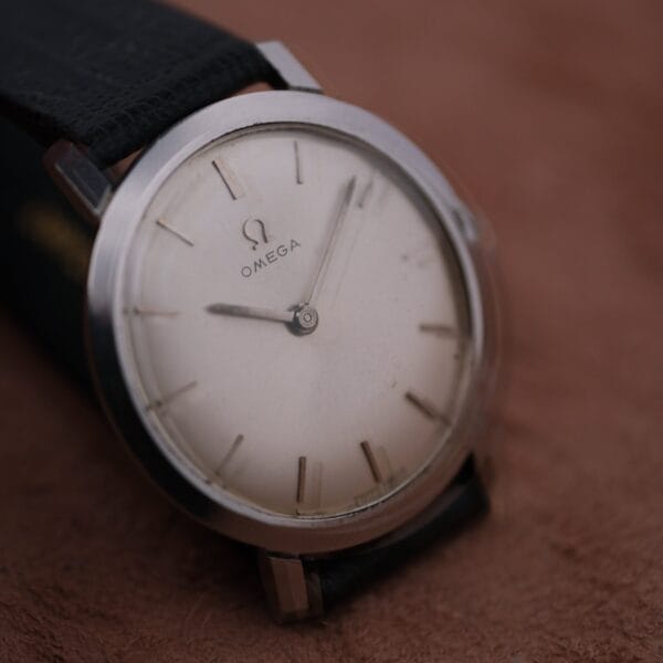 A close-up of the 1962 Omega Slim Dress Sunburst Dial wristwatch featuring a minimalist white dial and silver hour markers, paired with a black leather strap, is placed on a brown surface.