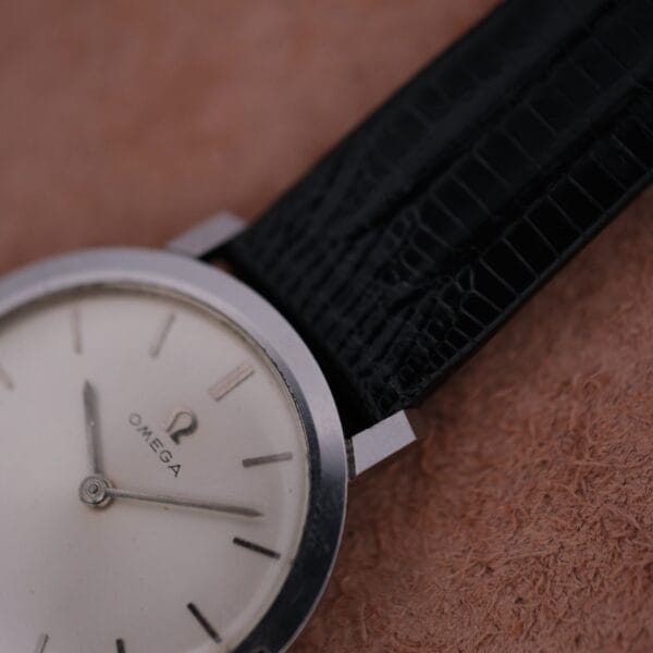 Close-up of an Omega 1962 Slim Dress Sunburst Dial on a leather strap, featuring a silver bezel and simple white face with slim hour markers and one visible hand against a textured brown background.