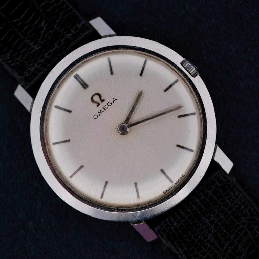 The 1962 Omega Slim Dress Sunburst Dial features a striking sunburst dial and black leather strap, beautifully presented against a dark background.