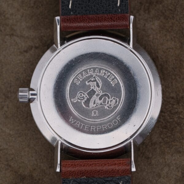A close-up of the 1963 Omega Seamaster Deville Türler Dial Ref. 135.010’s caseback, featuring an engraved seahorse emblem, SEAMASTER, WATERPROOF, and the Omega logo. The watch is paired with a black and brown leather strap.