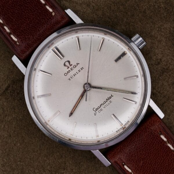 A 1963 Omega Seamaster Deville Türler Dial Ref. 135.010 wristwatch with a brown leather strap rests on a dark textured surface.