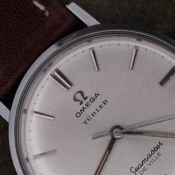 Close-up of a 1963 Omega Seamaster Deville Türler Dial Ref. 135.010 wristwatch with a silver dial, TÜRLER branding, black hour markers, and a brown leather strap partly visible. The time reads about 8:12:33.