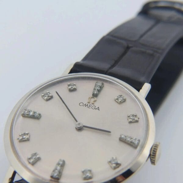 A close-up of the 1965 Omega Deville Ref. D6672 wristwatch features a silver dial with diamond hour markers, a black leather strap, and thin silver hands. The Omega logo is visible below the 12 oclock position.