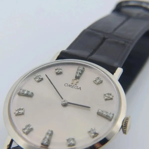A close-up of the 1965 Omega Deville Ref. D6672 wristwatch features a silver dial with diamond hour markers, a black leather strap, and thin silver hands. The Omega logo is visible below the 12 oclock position.