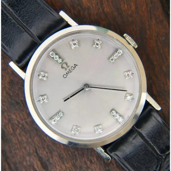 The 1965 Omega Deville 14k Ref. D6672 wristwatch, with silver hour markers and diamond accents, is displayed on a wooden surface. It features a round face, a black leather strap, and two black hands for hours and minutes.