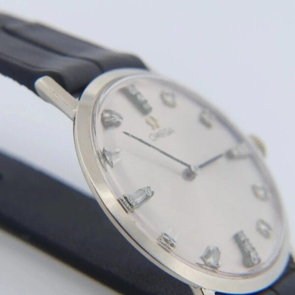 Close-up of a 1965 Omega Deville Ref. D6672 wristwatch featuring a silver case, black leather strap, white dial, and silver hour markers with thin hands. Classic and minimalist in design.