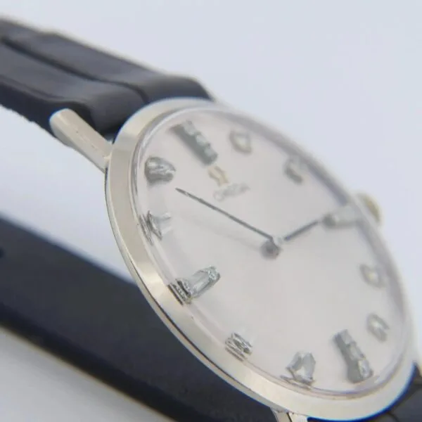Close-up of a 1965 Omega Deville Ref. D6672 wristwatch featuring a silver case, black leather strap, white dial, and silver hour markers with thin hands. Classic and minimalist in design.