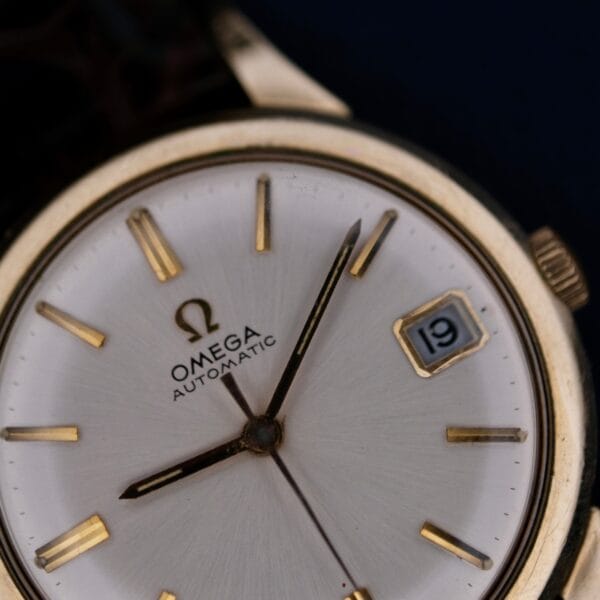 A close-up of the 1966 Omega Automatic Date Cal. 563 wristwatch features gold hour markers, gold hands, a date window showing 19, and the Omega logo on a silver dial. The time displayed is about 2:36.