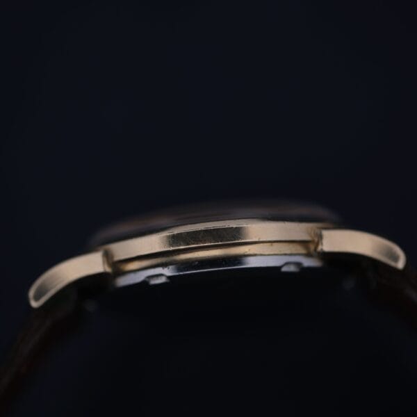 Close-up side view of the 1966 Omega Automatic Date Cal. 563 wristwatch case, highlighting its thin gold-colored metal profile against a dark background, with parts of the strap and buttons visible.