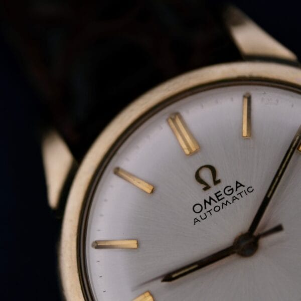 Close-up of a 1966 Omega Automatic Date Cal. 563 wristwatch with a silver dial, gold hour markers, and gold hands. The Omega logo and “Automatic” are visible on the watch face.