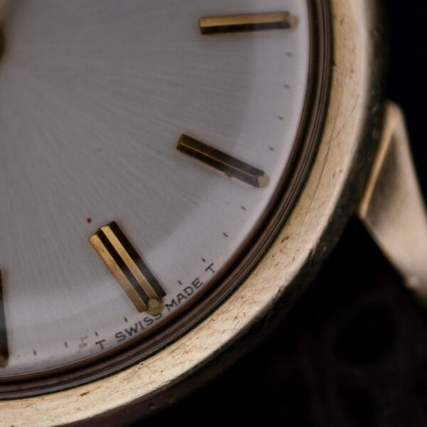 A close-up of the 1966 Omega Automatic Date Cal. 563 watch face displays gold hour markers, part of the bezel, and SWISS MADE near the bottom, focusing on the area between the 5 and 6 oclock markers.