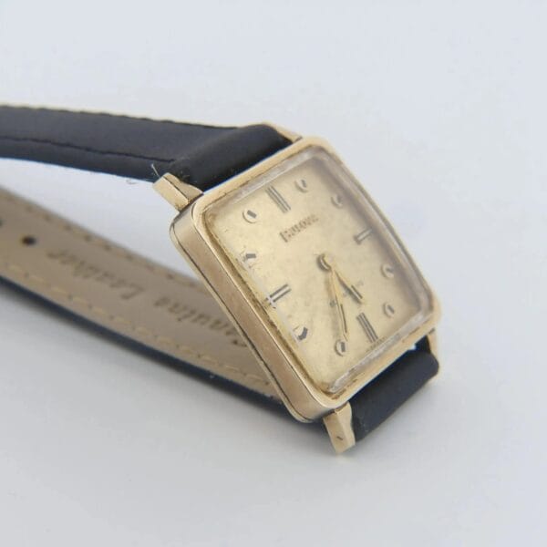 The 1968 BULOVA Automatic ‘Edwardian’ Vintage Square Dress Watch, featuring a black leather strap and gold case, is displayed against a light gray background. The beige dial with Roman numerals and slender black hands is slightly tilted for emphasis.