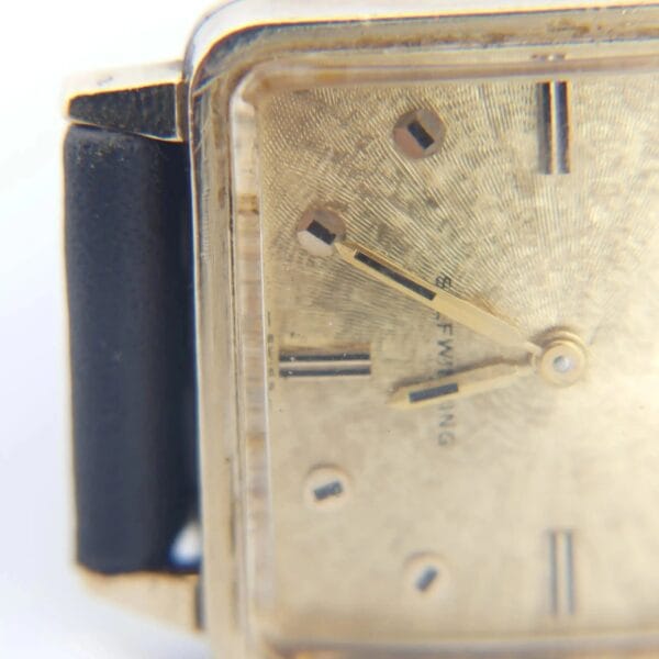 Close-up of the 1968 BULOVA Automatic ‘Edwardian’ Vintage Square Dress Watch, featuring a textured gold face, black hour markers, and a black leather strap. The watch hands show about 10:10 against a soft white blurred background.