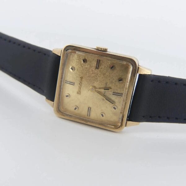 A 1968 BULOVA Automatic Edwardian vintage square dress watch with a gold finish and black leather strap is showcased. It features simple hour markers and two hands, showing some wear with minor scratches on the gold.