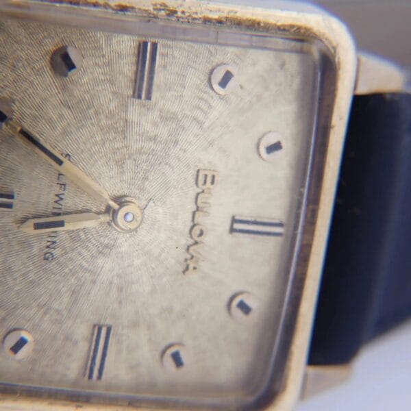 Close-up of a 1968 BULOVA Automatic Edwardian vintage square watch with a gold bezel, textured silver dial, and black leather strap. It features simple hour markers, small metallic hands, and a slightly worn face showing signs of age.