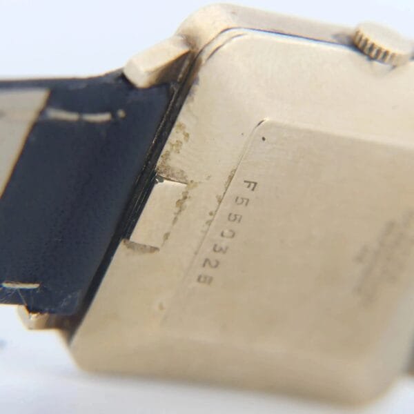 Close-up image of a slightly tarnished gold 1968 BULOVA Automatic Edwardian Vintage Square Dress Watch with a black strap. The watch case is inscribed with the serial number F55032B, highlighting the serial number and strap attachment.