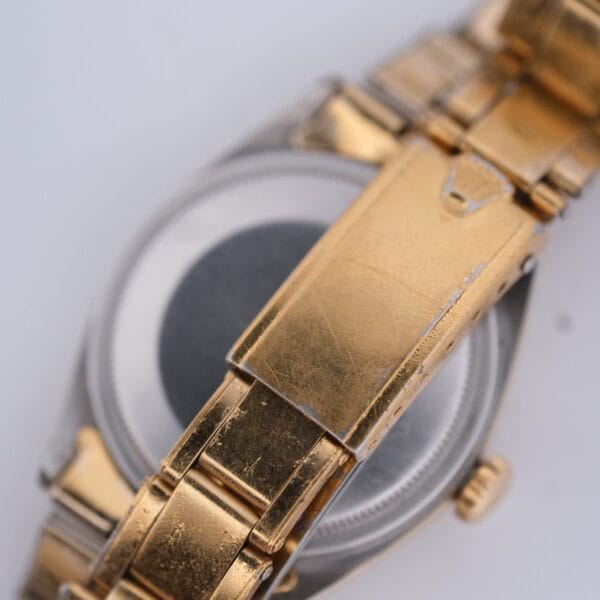 A close-up of a 1974 Rolex Oyster Perpetual Date Ref. 1550 with a gold and silver band shows the clasp featuring scratches and signs of wear. The circular metal watch case is partially visible beneath the band.