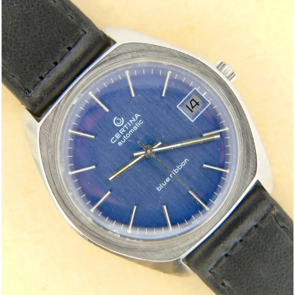 A 1976 Certina Blue Ribbon Automatic for Volvo featuring a blue dial, silver hour markers, date at 3 o’clock, and blue ribbon text, set in a silver case with a black leather strap.