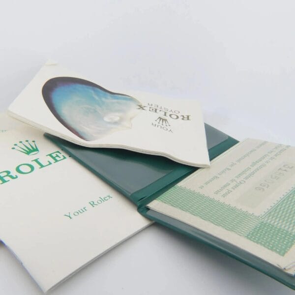 A green holder containing a Rolex certificate card, an open booklet with a pearl oyster image, and a document featuring text, with the Rolex logo and Your Rolex partially visible. Product: 1978 Rolex 1550 Oyster Perpetual Date 14k Gold Shell.