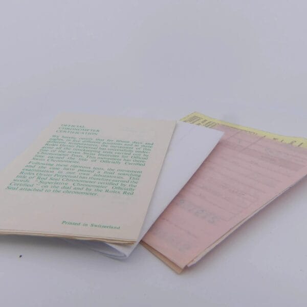 A small stack of different colored papers lies on a white surface, the top page with green text reading Official Certificate. The papers are slightly askew, revealing parts of each. The bottom reads Printed in Switzerland, evoking the elegance of a 1978 Rolex 1550 Oyster Perpetual Date 14k Gold Shell.
