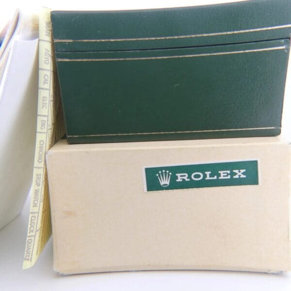 A beige box labeled ROLEX in green sits beside a green folder. Visible behind the folder are papers or manuals, with tabs AUTOMATIC, GMT, and CHRONO, related to the 1978 Rolex 1550 Oyster Perpetual Date 14k Gold Shell.