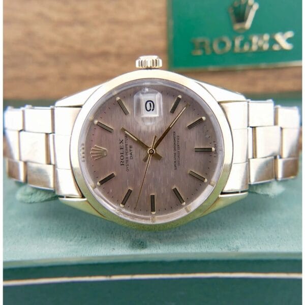A 1978 Rolex 1550 Oyster Perpetual Date with a metal band is shown on a green surface. The watch features a gray face, silver hour markers, and displays the date. The Rolex logo appears on a box in the background.