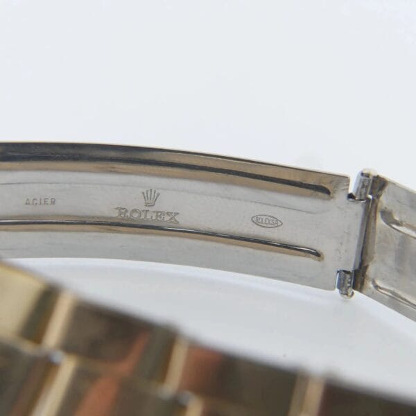 Close-up of the clasp inside a metal watch bracelet from a 1978 Rolex 1550 Oyster Perpetual Date 14k Gold Shell. It features engraved text ACIER and ROLEX with the iconic Rolex crown logo in between, while ROLEX appears in an oval, all on a shiny metal surface.