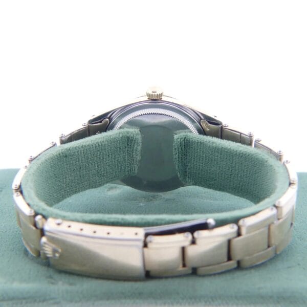 A 1978 Rolex 1550 Oyster Perpetual Date with a silver link band rests on a light green cushion. It is positioned to show the back and clasp, highlighting the polished finish and brand logo. The focus is on the rear of this 14k Gold Shell watch.