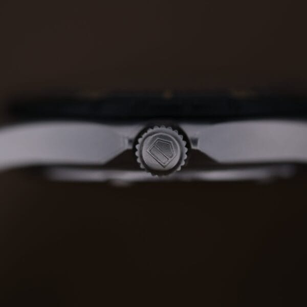 Close-up side view of the crown on a 1986 Tag Heuer 2000 Series Blue Bezel Ref. 972.606 1st Gen, featuring a shield-shaped logo engraving on its metallic case against a blurred brown background.