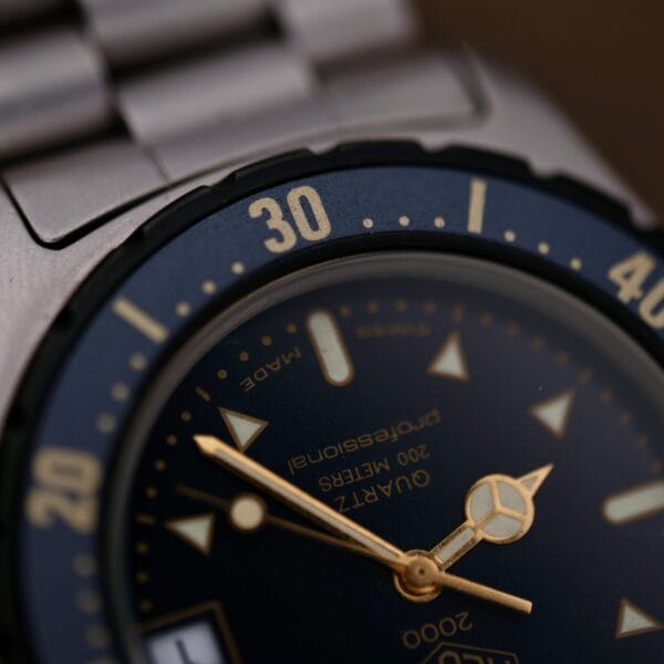 Close-up of a 1986 Tag Heuer 2000 Series Blue Bezel Ref. 972.606, showing part of the metal band, blue bezel with yellow numerals, visible hands and markers, and text reading Quartz, 200 meters, Professional.