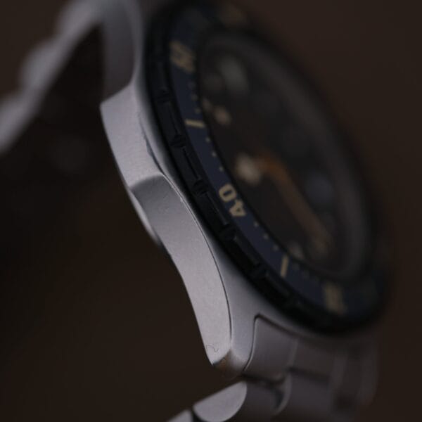 Close-up of the 1986 Tag Heuer 2000 Series Blue Bezel Ref. 972.606 (1st Gen) wristwatch, highlighting its metallic case and part of the blue bezel with white numbers; the watch face is blurred against a dark background.